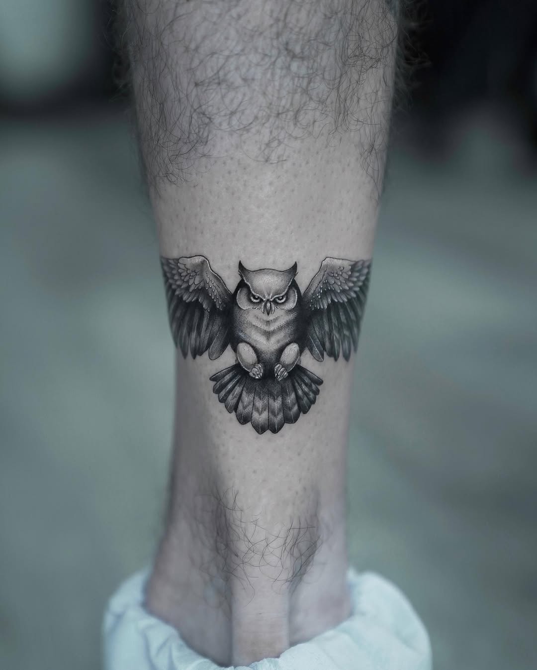 Black and grey realism owl tattoo on lower leg, detailed wings spread design with realistic shading and feather texture.
